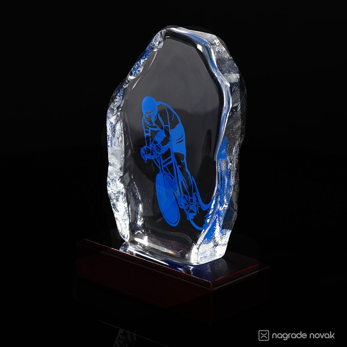 Cast glass award with a blue cyclist graphic and rough, ice-like edge, seen at an angle on a dark red wooden base