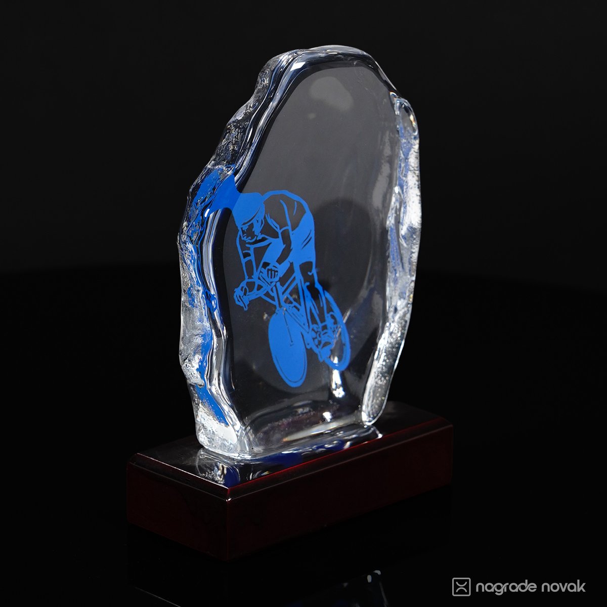 Side view of the cast glass award showing its thick, relief-textured edge, blue cyclist motif and dark wooden base