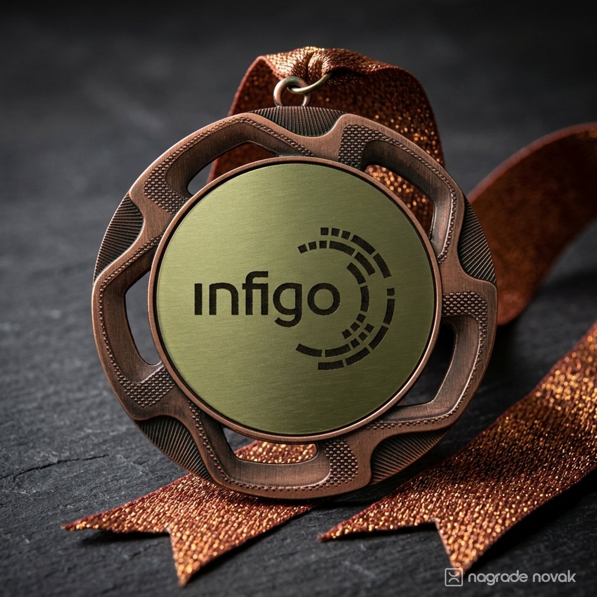 Close-up of a bronze medal with the black Infigo logo on a gold insert, resting on a dark slate surface with copper glitter ribbons