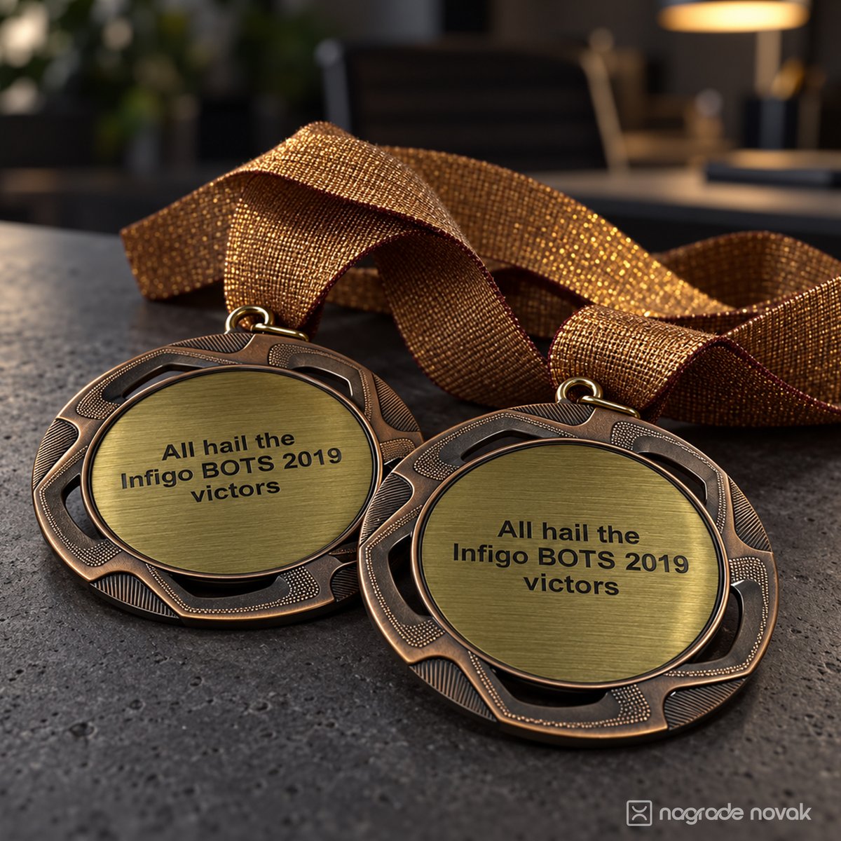 Two bronze medals with gold inserts reading "All hail the Infigo BOTS 2019 victors", on a dark stone surface with a gold glitter ribbon and a blurred office desk with a lamp in the background