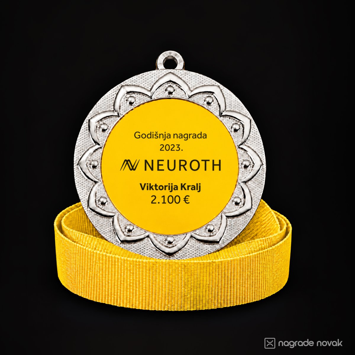 Silver medal with a petal-shaped embossed border and a yellow insert reading "Godišnja nagrada 2023." with the Neuroth logo and a personalised name and amount, standing on a coiled yellow ribbon on a dark background