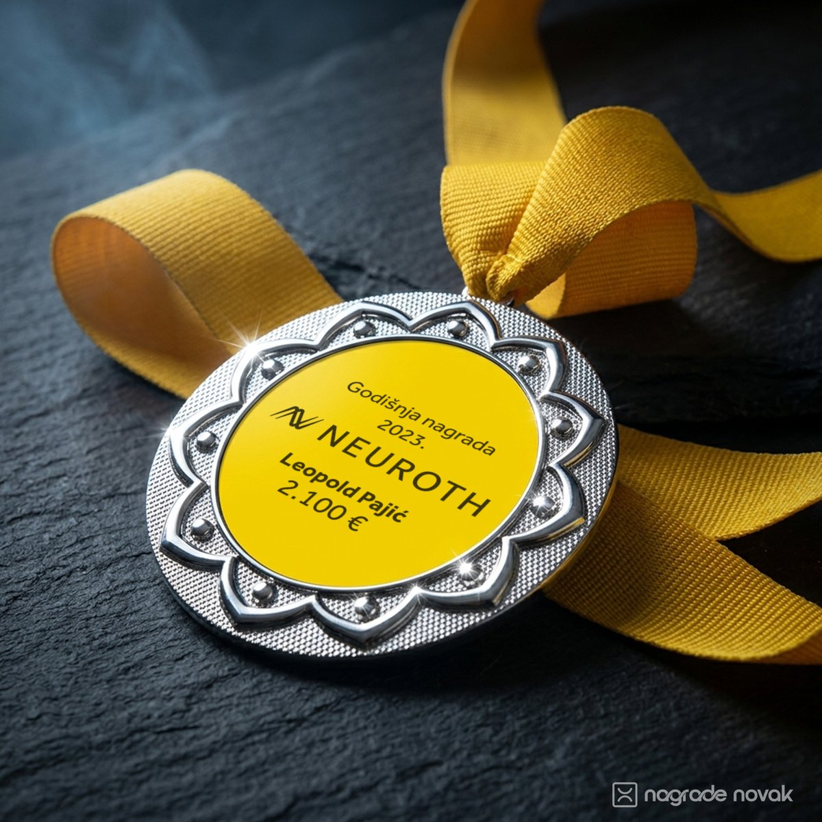 Silver medal with a yellow Neuroth insert, lying at an angle on dark slate with a yellow ribbon looped above it