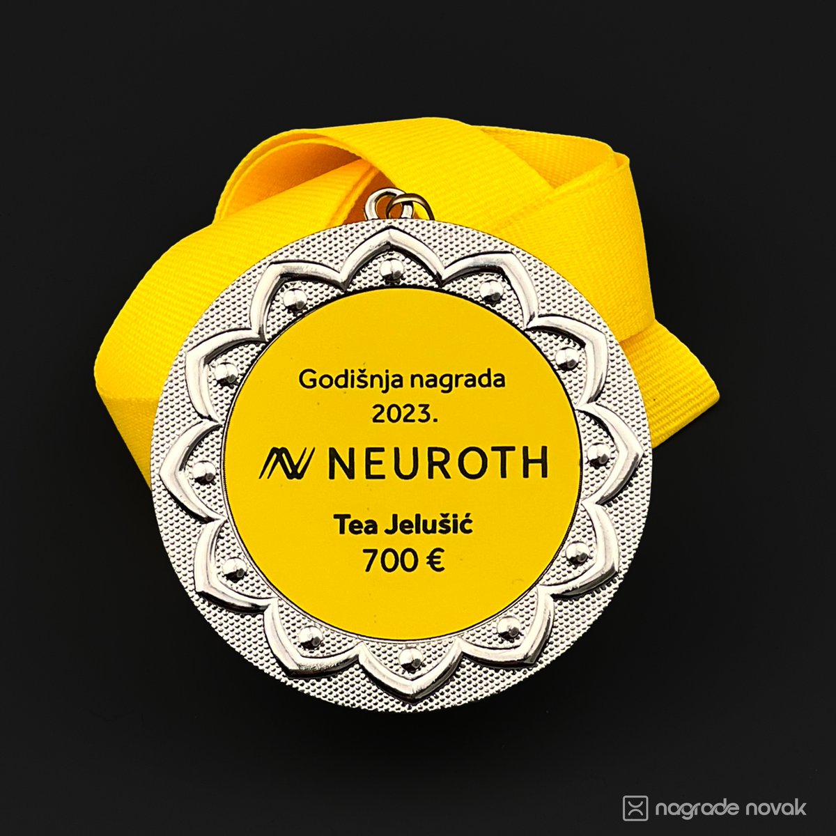 Close-up of a silver medal with a yellow insert reading "Godišnja nagrada 2023." with the Neuroth logo, a name and "700 €", with a yellow ribbon behind it