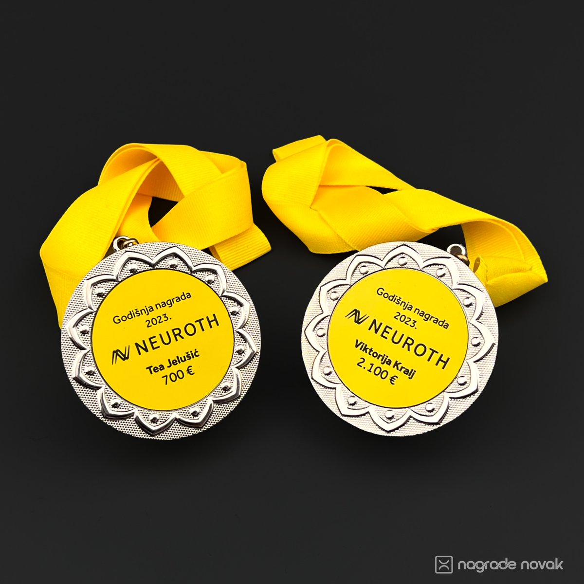 Two silver medals with yellow Neuroth inserts, each hanging from a yellow ribbon, lying side by side on a dark surface