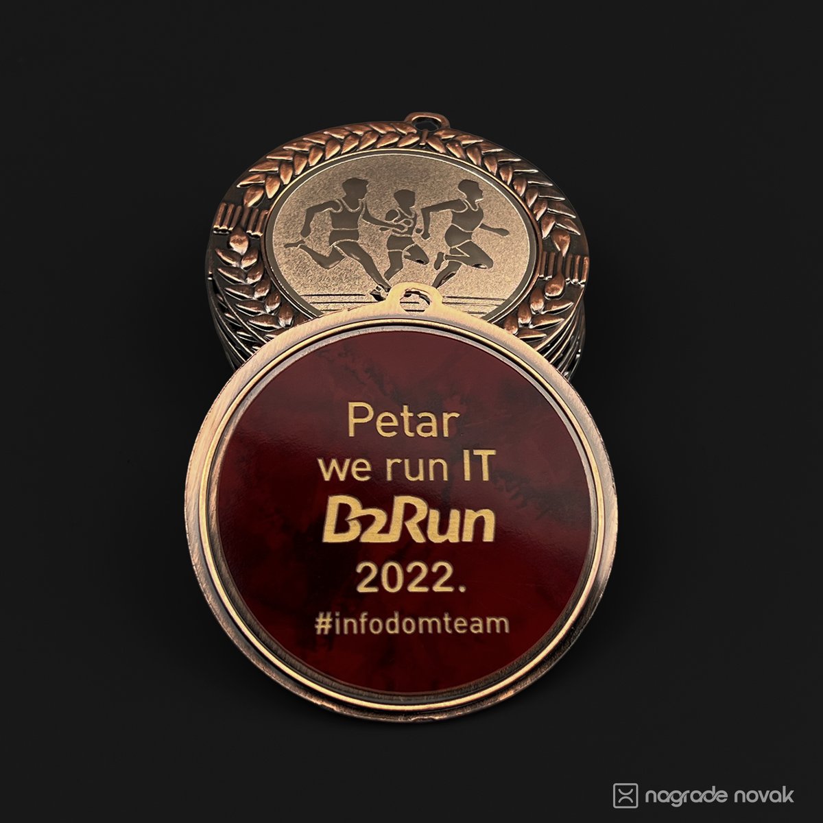 Close-up of a maroon and gold medal back insert reading “Petar, we run IT B2Run 2022”, in front of a stack of bronze medals with runners