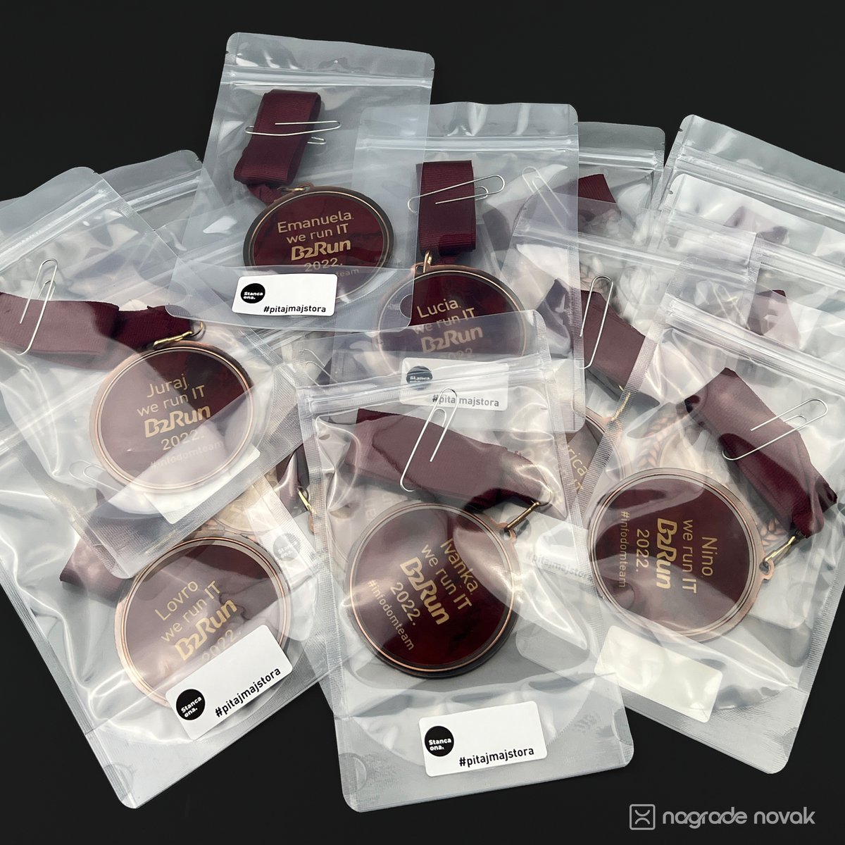 Pile of individually packed medals in clear zip bags with paper-clipped maroon ribbons and Stancaona labels, each personalised with a participant’s name, on a dark background