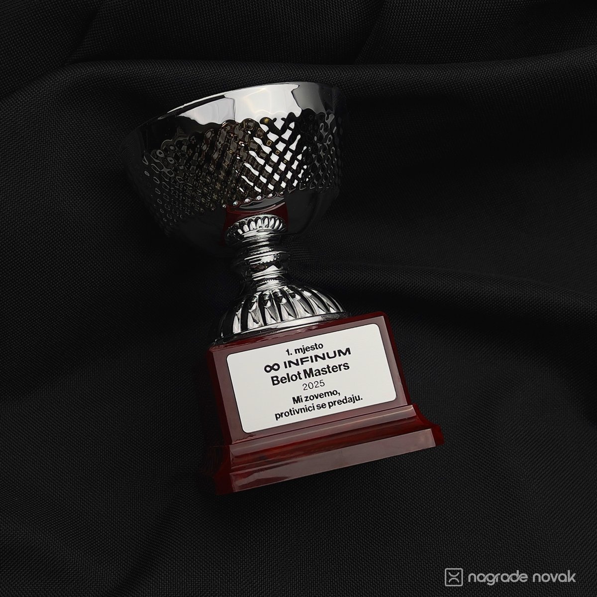 Angled shot of a silver trophy cup with a textured bowl on a dark red base, placed on dark fabric.