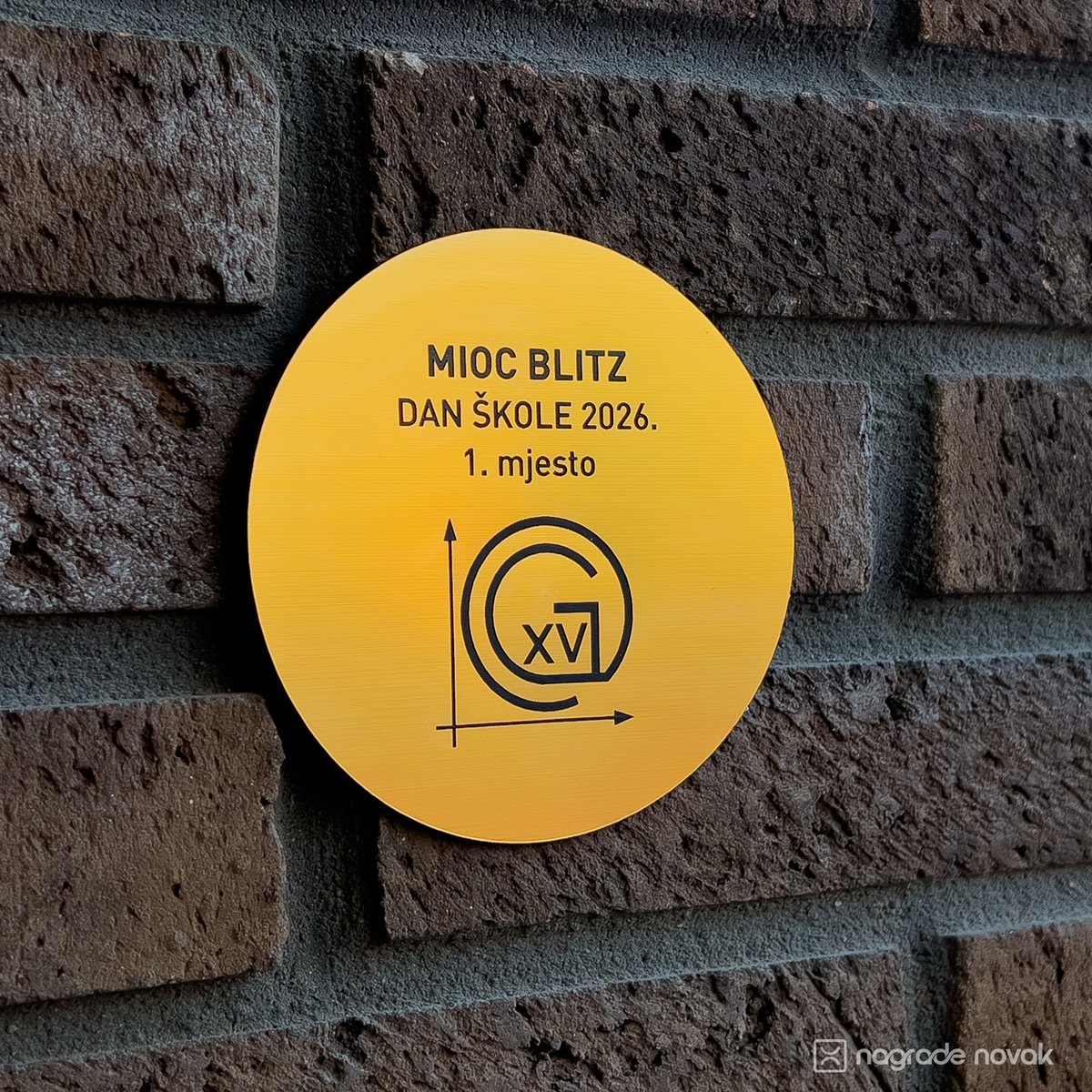 Round yellow gold plate with the text "MIOC BLITZ, DAN ŠKOLE 2026., 1. mjesto" and the school logo, mounted on a dark brick wall
