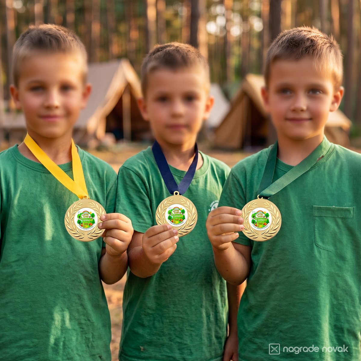 Three children in green T-shirts each showing a gold medal on a yellow, dark blue or green ribbon, in front of camp tents