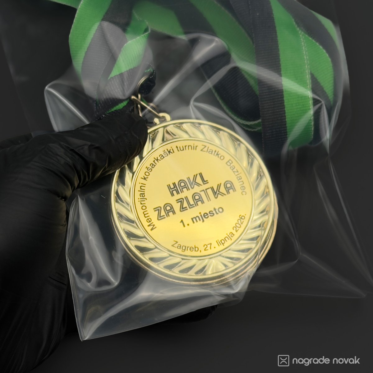 First-person view of a gloved hand holding a gold 1st place medal packed in a clear plastic bag with a green and black ribbon